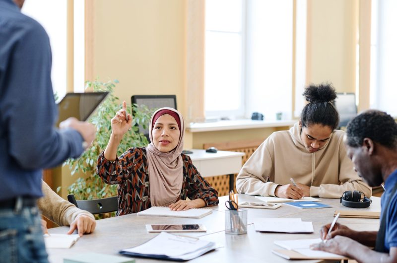 Muslim woman attending language lessons for immigrants raising hand to ask teacher question about task