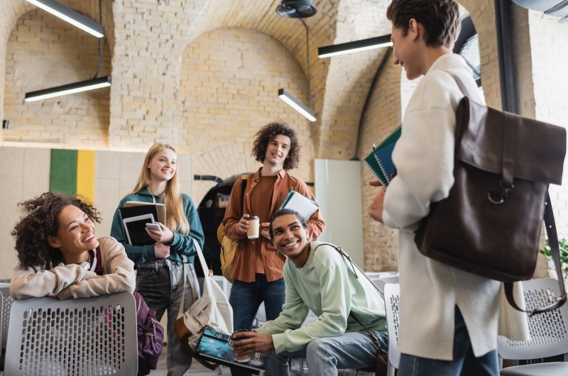 cheerful interracial classmates with gadgets and notebooks looking at student with leather  backpack on blurred foreground,stock image