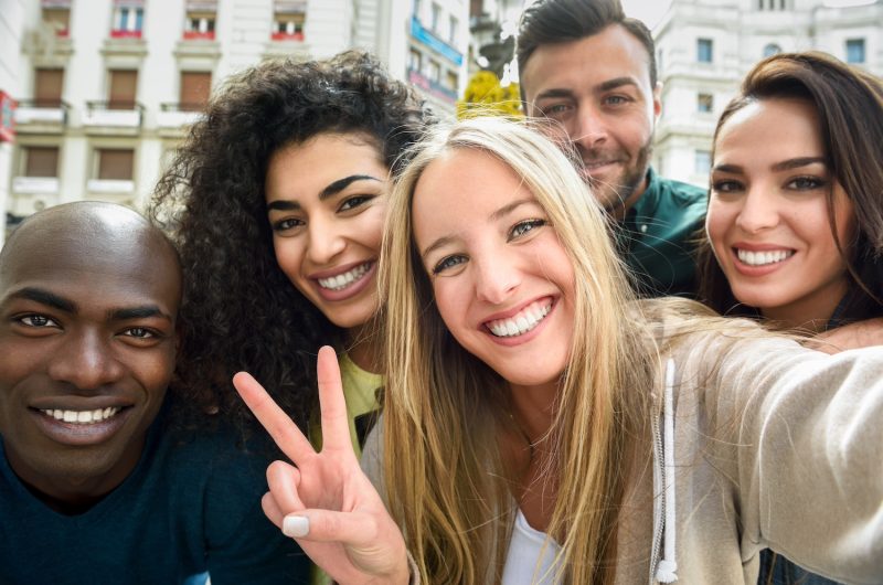 Multiracial group of friends taking selfie in a urban street with a blonde woman in foreground. Three young women and two men wearing casual clothes.